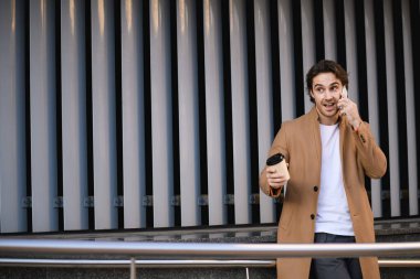 Kahverengi ceketli neşeli iş adamı akıllı telefonla konuşuyor ve elinde kağıt bardakla binanın yakınına geliyor.