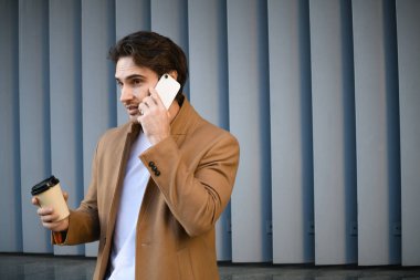 Ceketli girişimci cep telefonuyla konuşuyor ve dışarıda tek kullanımlık bardakları tutuyor.