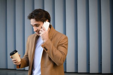 Pardösülü esmer işadamı elinde içecek ve dışarıda akıllı telefondan konuşuyor.
