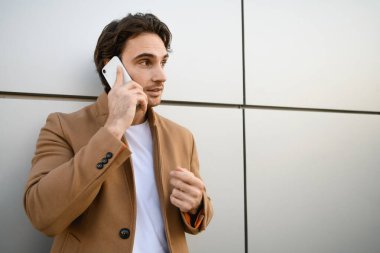 Palto giymiş yakışıklı bir adam binanın yakınındaki akıllı telefondan konuşuyor.