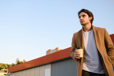 Sonbahar montlu yakışıklı adamın açık havada mavi gökyüzüne karşı içkisini yudumlarken alçak açılı görüntüsü.