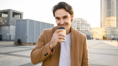 Kahverengi ceketli esmer adam elinde paket servis içkisi ve şehirde akşam vakti kameraya gülümsüyor.