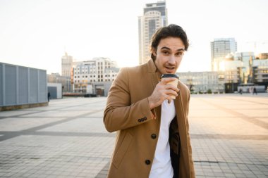 Ceketli yakışıklı bir adam kameraya bakıyor ve akşamları elinde kahve tutuyor.