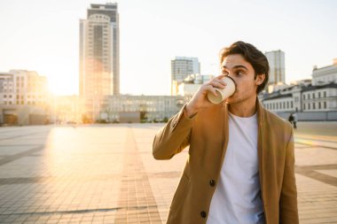 Paltolu ve beyaz tişörtlü esmer adam şehirde gün batımına karşı kahve içiyor.