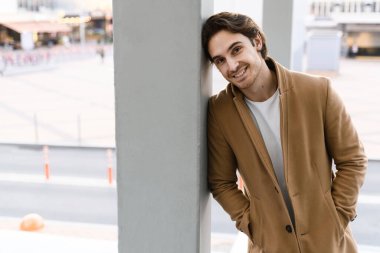 Kahverengi ceketli yakışıklı adam gülümsüyor ve binanın yakınındaki kameraya bakıyor.