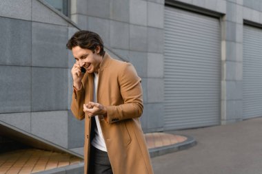 Kahverengi ceketli gülümseyen işadamı bulanık binanın yakınında akıllı telefondan konuşuyor.