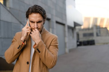 Kahverengi ceketli düşünceli bir işadamı şehir caddesinde cep telefonuyla konuşuyor.