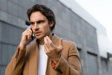 Paltolu stresli iş adamı. Gündüz vakti akıllı telefondan konuşuyor.