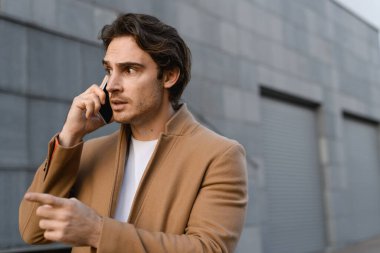 Palto giymiş bir işadamı akıllı telefondan konuşuyor ve şehir caddesini işaret ediyor.