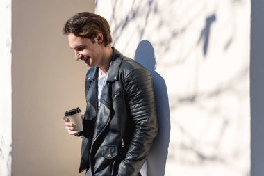 Deri ceketli, kahveli, gölgeli duvarın yanına yaklaşan olumlu adam.