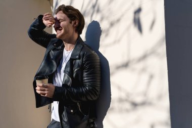 Deri ceketli mutlu adam güneşli sokakta kağıt bardak tutuyor.