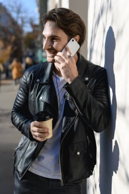 Deri ceketli gülümseyen adam akıllı telefondan konuşuyor ve elinde yemek kupası tutuyor.