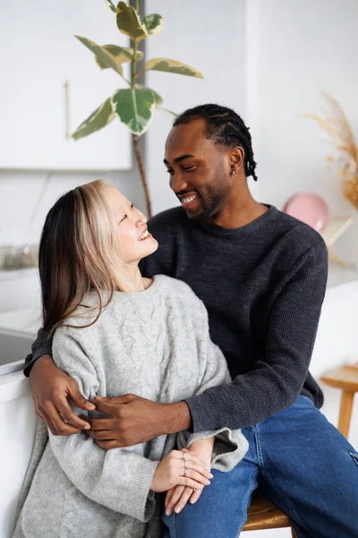 Gülümseyen Afrikalı Amerikalı genç adam modern mutfakta Asyalı kız arkadaşına sarılıyor.