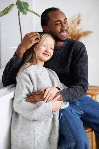 Gülümseyen Afrikalı Amerikalı genç adam mutfakta Asyalı kız arkadaşına sarılıyor.