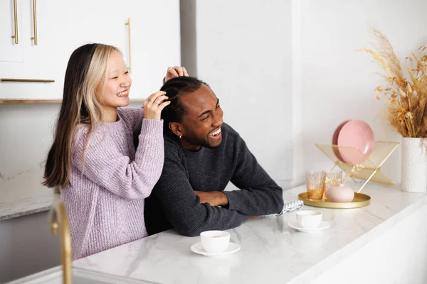 Süveter giyen genç Asyalı bir kadın, gülümseyen afro-amerikan erkek arkadaşının örgülerine dokunuyor.