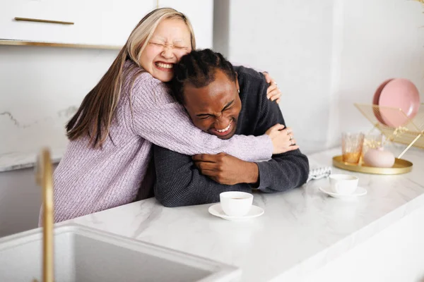 Mutlu genç Asyalı kadın, sabah kahvesinin yanında neşeli Afrikalı Amerikalı erkek arkadaşına sarılıyor.