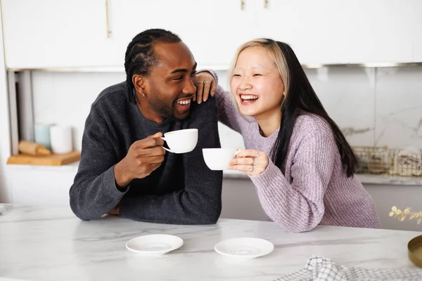 Evde mutfakta konuşurken kahve fincanı tutan pozitif ırklar arası genç bir çift.