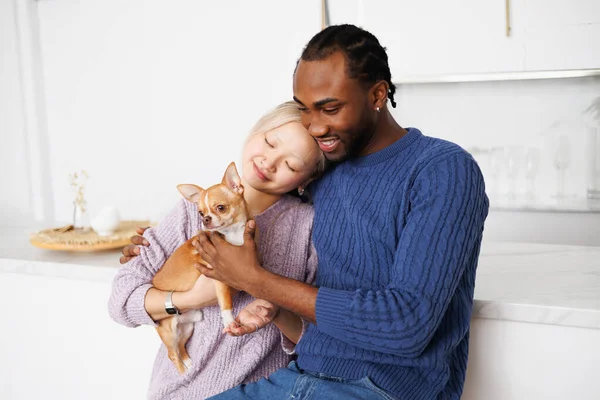 Süveter giyen, gülümseyen ve evde köpeğini tutan çok ırklı genç bir çift.