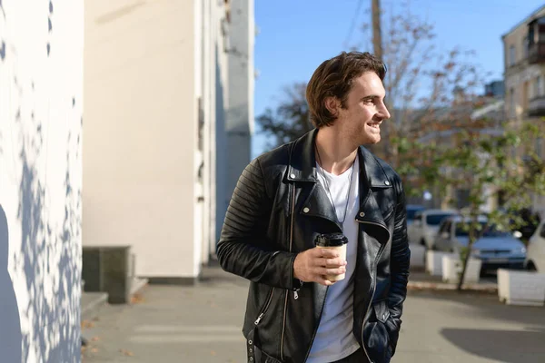 Deri ceketli yakışıklı adam şehirde sokağa çıkmak için kahve tutuyor.