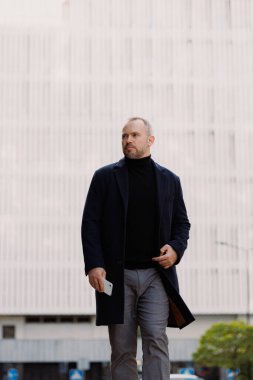 Palto ve balıkçı yakalı şık bir adam sokakta yürürken elinde akıllı telefon tutuyor.