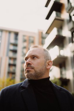 Balıkçı yaka ve palto giymiş yetişkin bir adamın şehir sokağına bakışları.