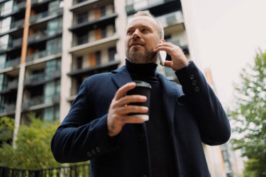 Zarif iş adamı akıllı telefondan konuşuyor ve sonbaharda şehir sokaklarında kahve içiyor.