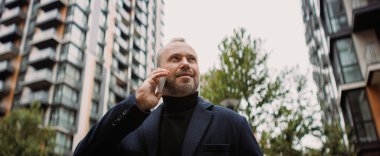 Palto giymiş gülümseyen bir işadamı şehirde akıllı telefondan konuşuyor.