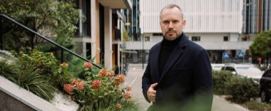 Paltolu yetişkin bir adam şehrin caddesindeki kameraya bakıyor.