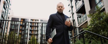 Ceketli şık bir işadamı kameraya bakıyor ve şehirde akıllı telefon ve dizüstü bilgisayar tutuyor.