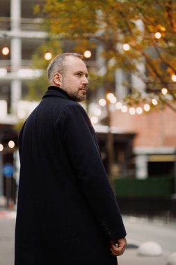 Palto giymiş sakallı adam sonbaharda şehir sokağına bakıyor.