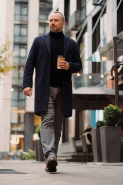 Paltolu ve elinde kahve tutan kendine güvenen bir adam sokakta yürürken.
