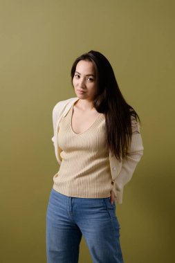 Esmer, hırkalı ve kot pantolonlu bir kadın yeşil, vücut pozitifliği olan kameraya bakıyor.