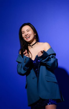 Neşeli ve şık Asyalı kadın ceketli ve mavi arka planda boğucu