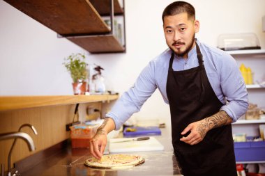 Asyalı şef pizzaya ananas koyuyor ve kafe mutfağındaki kameraya bakıyor.