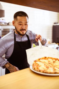 Önlüklü Asyalı şef kafede taze pişmiş pizzayı tabağa koyuyor.