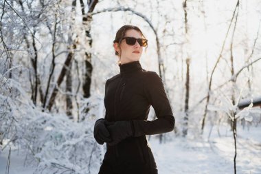 Aktif giyinen esmer kadın, güneş gözlüğü ve eldivenler karlı ormanda duruyor.