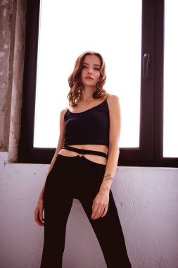 Evin penceresinin yanındaki kameraya bakan moda bir kadın.