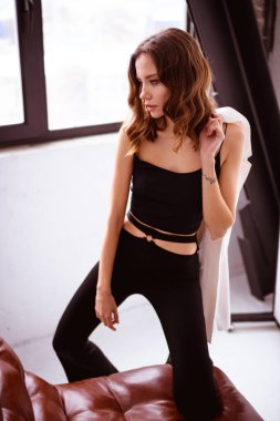 Ceket tutan ve evinde sahte deri koltuğun yanında poz veren moda bir kadın.