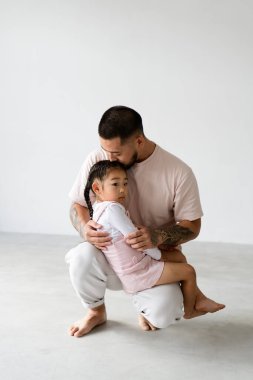 Genç dövmeli Asyalı baba, küçük kızını gri arka planda öpüyor.