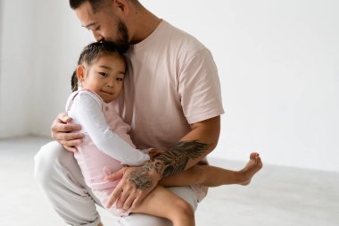 Asyalı baba, gri arka planda çıplak ayakla kızı öpüyor.