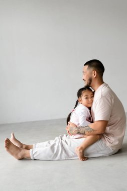 Gülümseyen Asyalı baba, gri arka planda otururken küçük kızını kucağına alıyor.