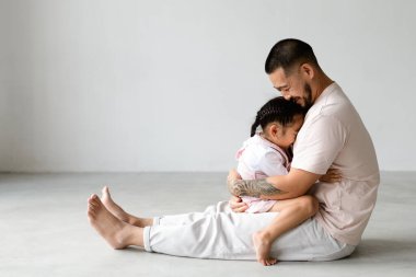 Yalınayak Asyalı baba neşeli küçük kızına sarılıyor ve stüdyoda gri arka planda eğleniyor.