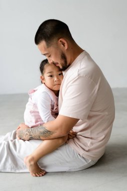 Küçük Asyalı çocuk gri arka planda babasının üzerine otururken kameraya bakıyor.