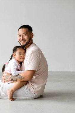 Neşeli Asyalı bir babanın portresi bebeğe sarılıyor ve gri arka planda kameraya bakıyor.