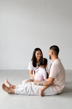 Küçük kızı olan modern Asyalı ebeveynler stüdyoda gri arka planda oturuyorlar.