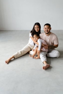 Olumlu genç Asyalı ebeveynler gri arka planda küçük kızlarını tutarken kameraya bakıyorlar.