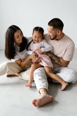 Genç, çıplak ayaklı Asyalı bir aile ve kızı gri arka planda otururken oynuyorlar.