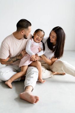 Gülümseyen genç Asyalı ebeveynler gri üzerinde otururken komik küçük kızlarıyla oynuyorlar.