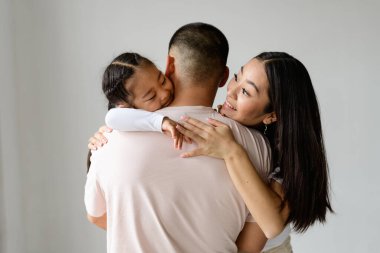 Gülümseyen Asyalı kadın yeni yürümeye başlayan kızıyla oynuyor ve sarılan koca griye bürünmüş.