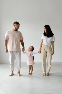 Genç Asyalı ebeveynler gri arka planda gülümseyen küçük kızın ellerini tutuyorlar.